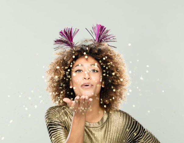 woman wearing gold blouse and party headband blowing sparkles.