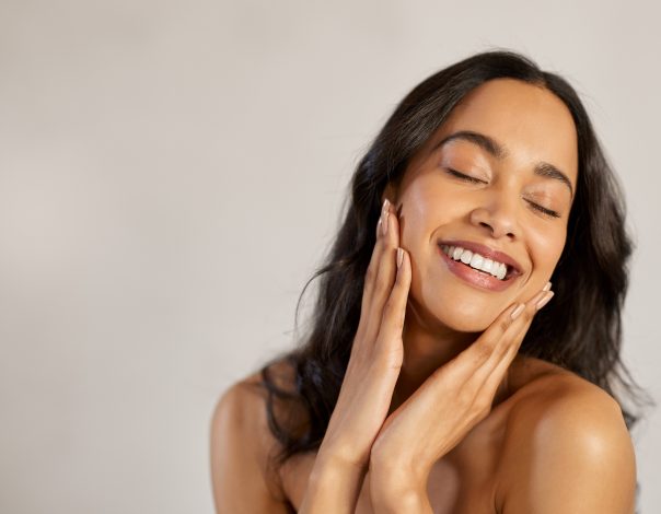 Portrait of beautiful latin woman touching her clean and healthy face against grey background. Smiling hispanic woman with natural makeup feeling healthy skin with eyes closed. Attractive multiethnic young woman pampering with perfect skin.